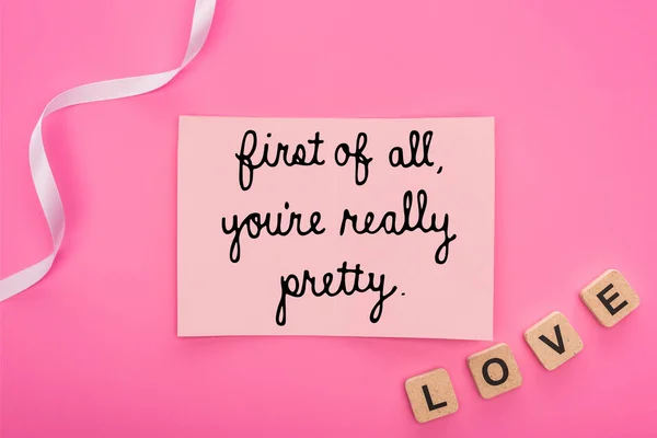 Top view of empty paper card with first of all you are really pretty lettering near ribbon and love lettering on wooden cubes isolated on pink — Stock Photo