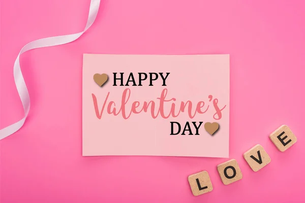 Top view of empty paper card with  happy valentines day lettering near ribbon and love lettering on wooden cubes isolated on pink — Stock Photo