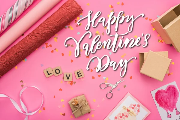 Top view of valentines confetti, scissors, wrapping paper, gift boxes, greeting cards and love lettering on wooden cubes on pink background with happy valentines day illustration — Stock Photo