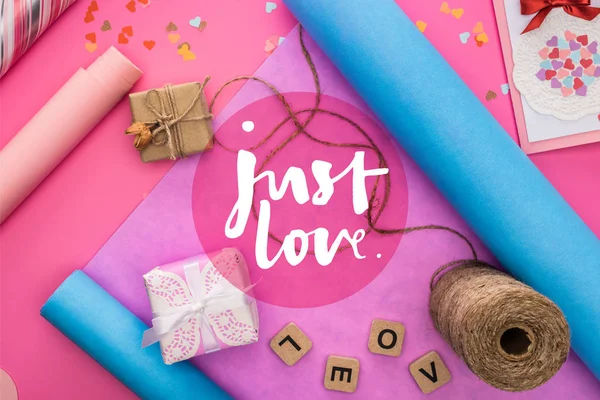 Top view of valentines decoration, wrapping paper, twine, gift boxes, greeting card and love lettering on wooden cubes on pink background with just love lettering — Stock Photo