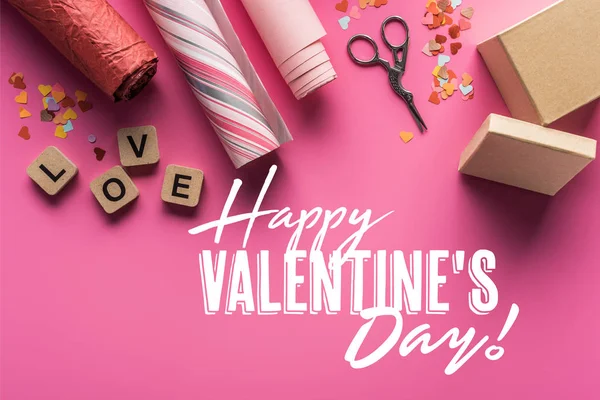 Ansicht von Valentinstag Dekoration, Schere, Geschenkschachtel, Geschenkpapier und Liebesschrift auf Holzwürfeln auf rosa Hintergrund mit glücklichen Valentinstag Illustration — Stockfoto