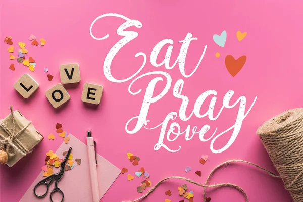 Top view of valentines decoration, scissors, gift box, twine and love lettering on wooden cubes on pink background with eat, pray, love lettering — Stock Photo