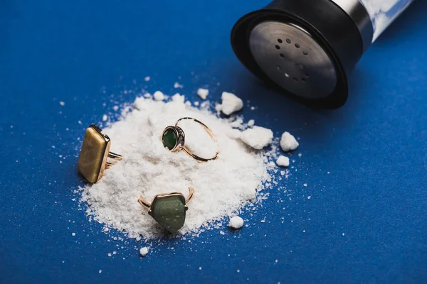 Close up view of jewellery rings on salt heap with salt shaker on blue surface — Stock Photo