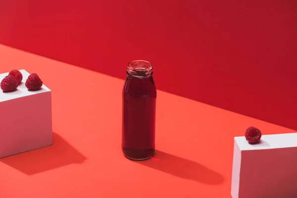 Frischer Beerensaft in Glasflasche in der Nähe reifer Himbeeren auf Würfeln auf rotem Hintergrund — Stockfoto