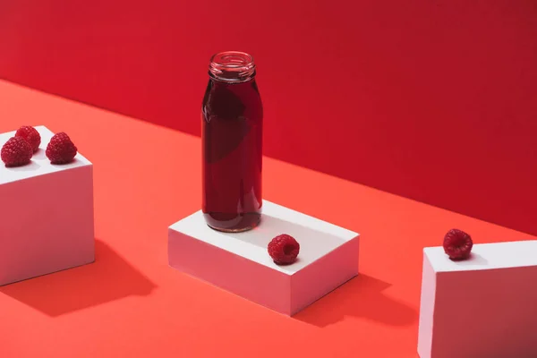 Jus de baies fraîches en bouteille de verre près de framboises mûres sur cubes sur fond rouge — Photo de stock