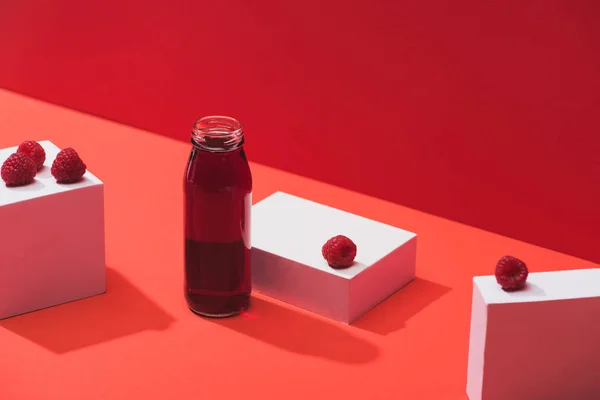 Jus de baies fraîches en bouteille de verre près de framboises mûres sur cubes sur fond rouge — Photo de stock