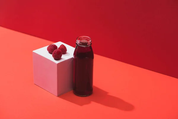 Jus de baies fraîches en bouteille de verre près de framboises mûres sur cube sur fond rouge — Photo de stock