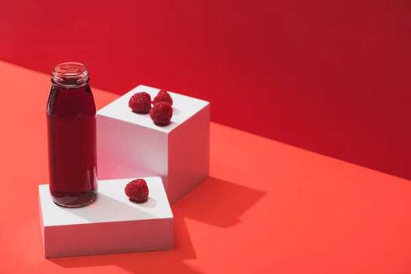 Jus de baies fraîches en bouteille de verre près de framboises mûres sur cubes sur fond rouge — Photo de stock