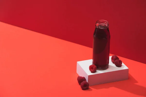 Succo di bacca fresco in bottiglia di vetro vicino a lamponi maturi su cubo su sfondo rosso — Foto stock