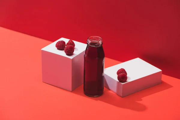 Свежий ягодный сок в стеклянной бутылке возле спелой малины на кубиках на красном фоне — стоковое фото