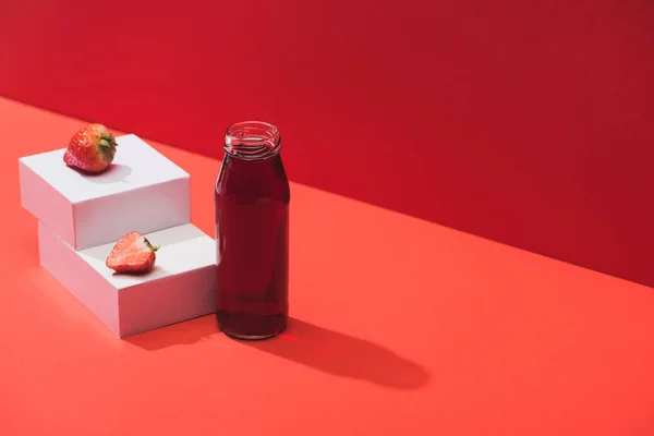 Suco de baga fresco em garrafa de vidro perto de morango maduro em cubos no fundo vermelho — Fotografia de Stock