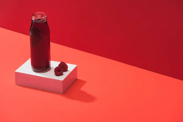 Frischer Beerensaft in Glasflasche in der Nähe reifer Himbeeren auf Würfel auf rotem Hintergrund — Stockfoto