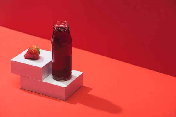 Fresh berry juice in glass bottle near ripe strawberry on cubes on red background — Stock Photo