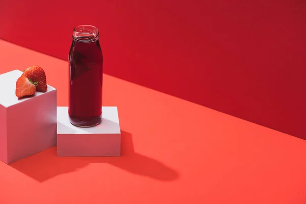 Fresh berry juice in glass bottle near ripe strawberry on cubes on red background — Stock Photo