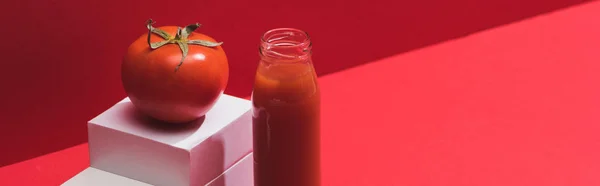 Свіжий овочевий сік у скляній пляшці біля стиглого помідора на підставці на червоному тлі, панорамний знімок — стокове фото