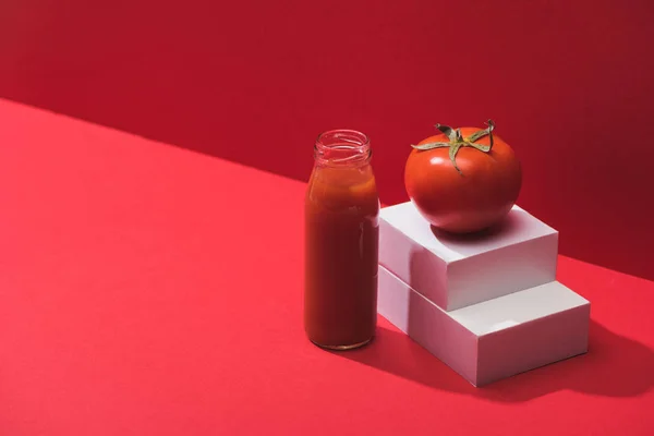 Свіжий овочевий сік у скляній пляшці біля стиглого помідора на стійці на червоному тлі — стокове фото