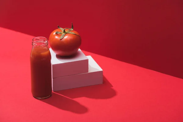 Fresh vegetable juice in glass bottle near ripe tomato on stand on red background — Stock Photo