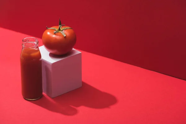 Fresh vegetable juice in glass bottle and ripe tomato on cube on red background — Stock Photo