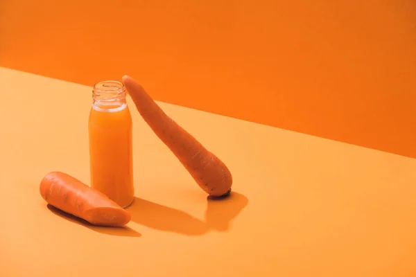 Fresh juice in glass bottle near ripe carrots on orange background — Stock Photo
