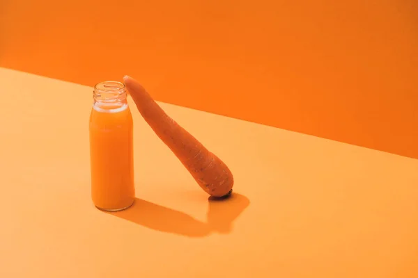 Jus frais en bouteille de verre près de carotte mûre sur fond orange — Photo de stock