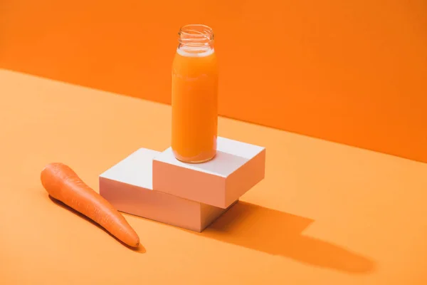 Fresh juice in glass bottle on cubes near ripe carrot on orange background — Stock Photo