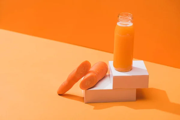 Fresh juice in glass bottle near ripe carrots on cubes on orange background — Stock Photo