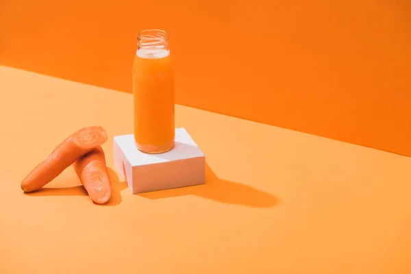 Fresh juice in glass bottle on cube near ripe carrots on orange background — Stock Photo