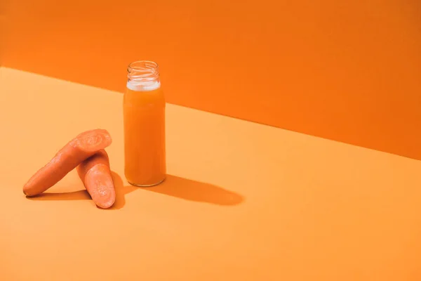 Свежий сок в стеклянной бутылке возле спелых морковок на оранжевом фоне — стоковое фото