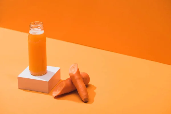 Fresh juice in glass bottle on cube near ripe carrots on orange background — Stock Photo