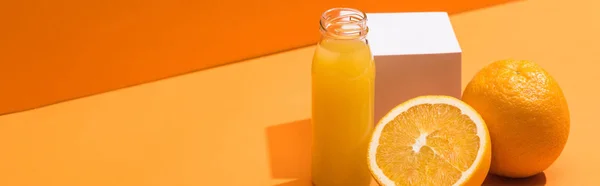 Fresh juice in glass bottle near oranges and white cube on orange background, panoramic shot — Stock Photo