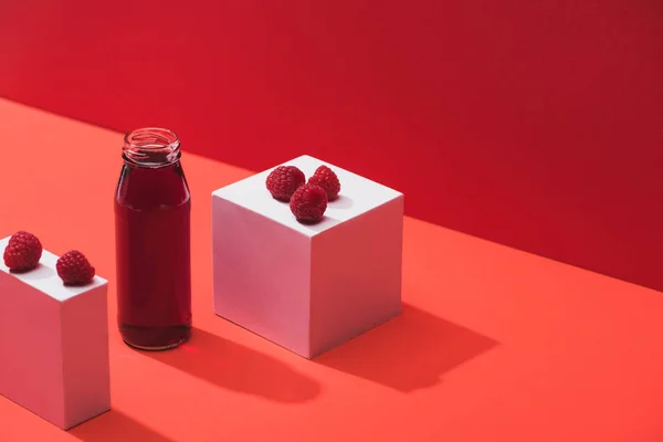 Jus de baies fraîches en bouteille de verre près de framboises mûres sur cubes sur fond rouge — Photo de stock