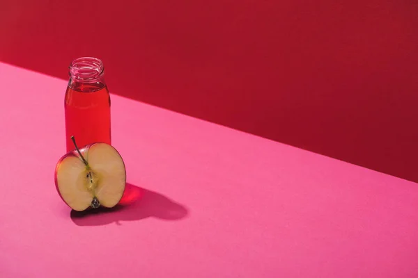 Frischer Saft in der Flasche in der Nähe der Apfelhälfte auf rotem und rosa Hintergrund — Stockfoto