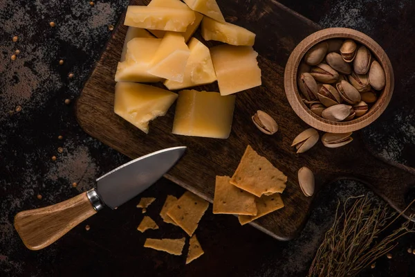 Vista superior de bandeja de queso con pistachos y galletas saladas sobre tabla de cortar de madera cerca de cuchillo - foto de stock