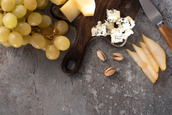 Top view of pieces of cheese on cutting board next to grapes, pistachios, knife and slices of pear on grey background — Stock Photo