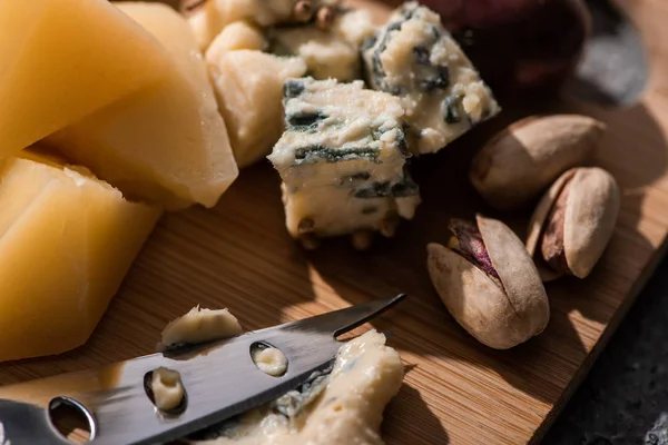 Selective focus of dorblu pressed by knife next to pieces of grana padano and pistachios on cutting board — Stock Photo