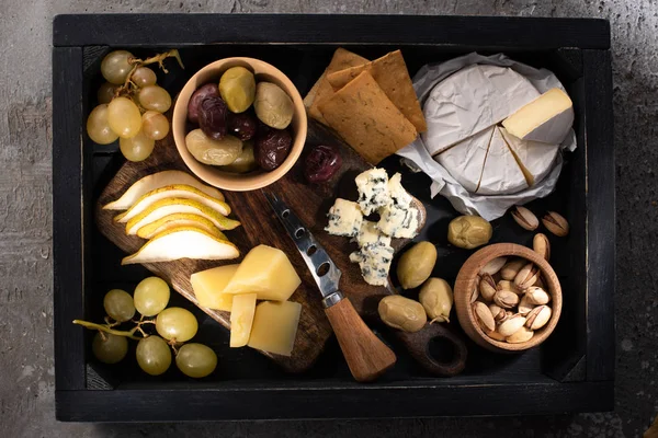 Top view of different kinds of cheese with fruits and nuts around knife on tray on grey background — Stock Photo