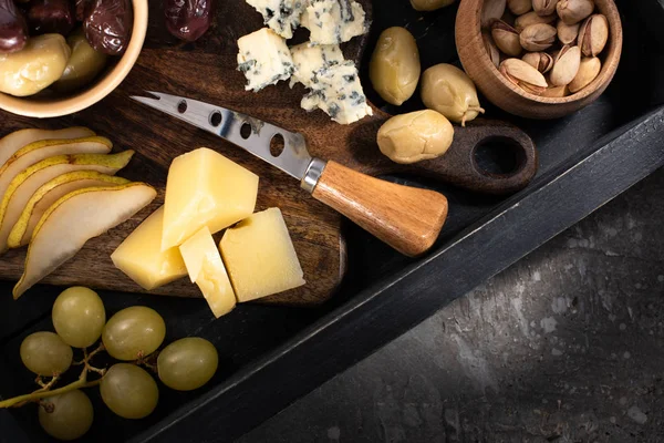 Top view of tray with pieces of cheese, dried olives, pistachios, slices of pear, grapes and knife on grey background — Stock Photo