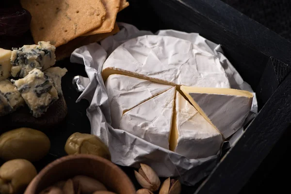 Selective focus of pieces of camembert next to dorblu, dried olives, crackers and pistachios on tray on black background — Stock Photo