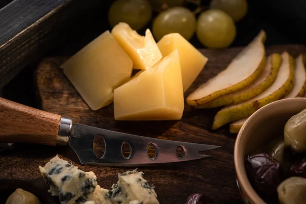 Selective focus of knife, pieces of cheese, slices of pear, dried olives, grapes on cutting board on tray — Stock Photo