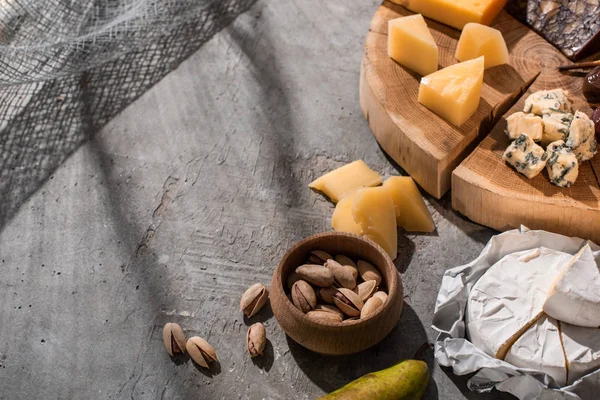 Vista en ángulo alto de pera, pistachos y camembert junto a diferentes tipos de queso sobre tabla de madera sobre fondo gris - foto de stock