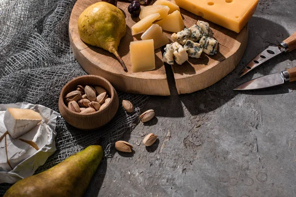 Trozos de queso con pera y aceitunas sobre tabla de madera junto a cuchillos, nueces sobre fondo gris - foto de stock