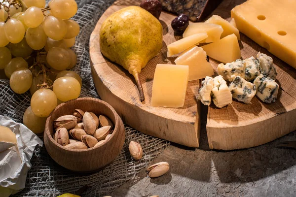Enfoque selectivo de trozos de queso con pera y aceitunas sobre tabla de madera junto a uvas y frutos secos sobre fondo gris - foto de stock