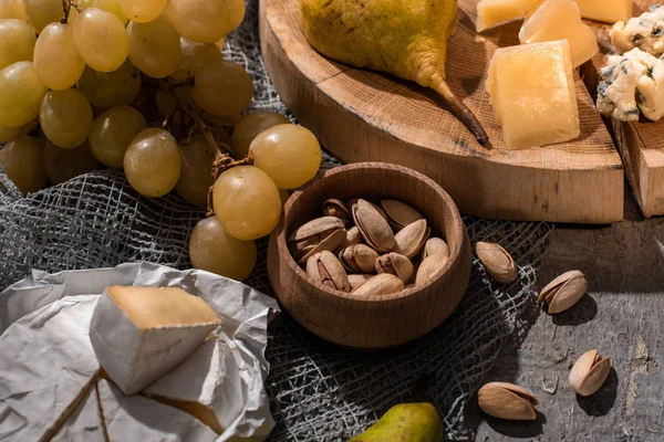 Cheese with pear on wooden board next to grapes, pistachios and Camembert on grey background — Stock Photo