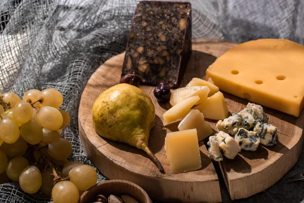 Selective focus of different kinds of cheese with pear and olives on wooden board next to grapes and pistachios on grey background — Stock Photo