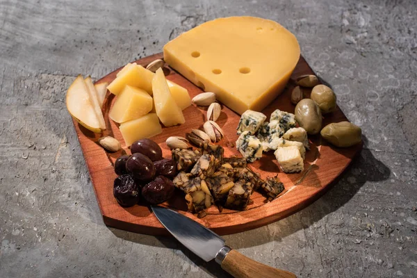 Composición alimenticia de queso con aceitunas secas, trozos de pera, pistachos y cuchillo sobre tabla de cortar sobre fondo gris - foto de stock
