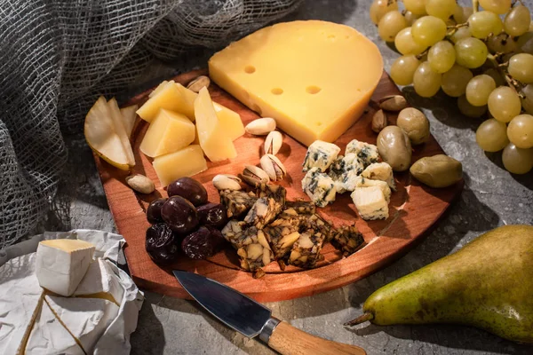 Pera, Camembert, uvas y cuchillo junto a tabla de cortar con composición de alimentos sobre fondo gris - foto de stock