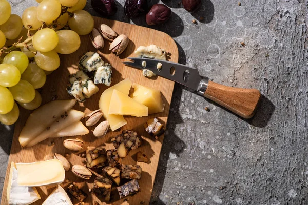 Top view of pieces of cheese, olives, grapes, pistachios, slices of pear and knife on cutting board on grey background — Stock Photo