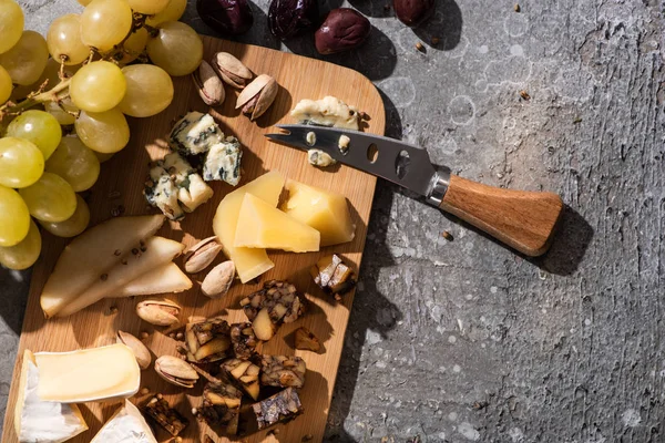 Vista superior de trozos de queso, aceitunas, uvas, pistachos, rodajas de pera y cuchillo sobre tabla de cortar sobre fondo gris — Stock Photo