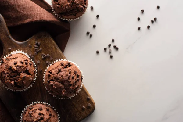 Vista superior de muffins de chocolate fresco na placa de corte de madeira perto de guardanapo marrom na superfície de mármore — Fotografia de Stock