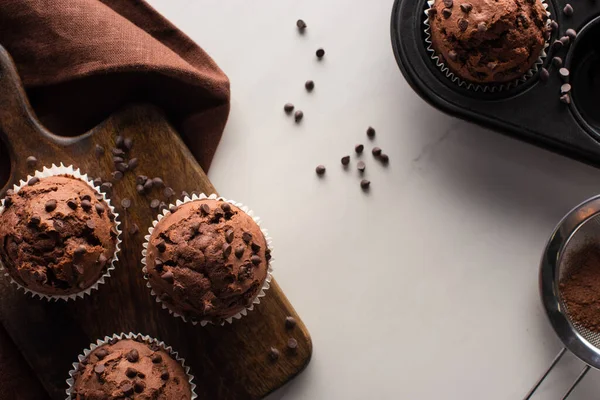 Vue du dessus des muffins au chocolat frais sur planche à découper en bois près de la serviette brune et de la poudre de cacao sur la surface du marbre — Photo de stock